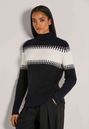 Frau mit langen dunklen Haaren, die einen schwarz-weißen gerippten Strickpullover und schwarze Hosen trägt, steht vor einer schlichten hellen Wand.