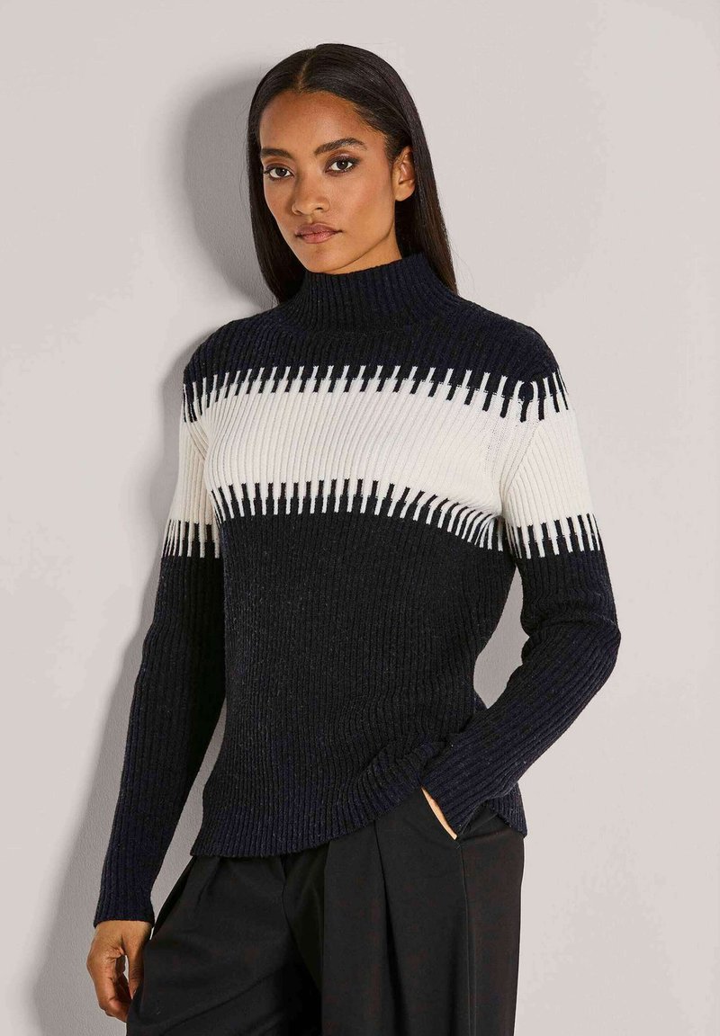Frau mit langen dunklen Haaren, die einen schwarz-weißen gerippten Strickpullover und schwarze Hosen trägt, steht vor einer schlichten hellen Wand.