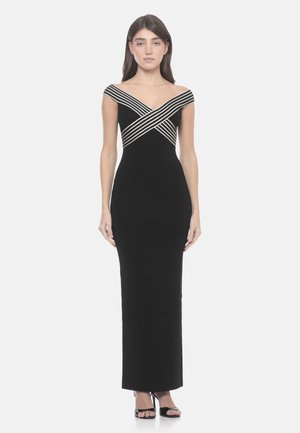 Donna in piedi che indossa un lungo vestito nero con dettaglio a righe bianche incrociate sulle spalle scoperte e sandali neri con tacco.