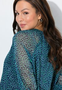 Průsvitná tmavě modrá halenka se zeleným vzorem ve tvaru srdce. Látka jemně splývá kolem ramen, s nařaseným výstřihem a volným střihem.