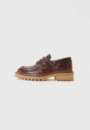 Mocassin en cuir marron à talon bas avec semelle en caoutchouc épaisse couleur clair et détails de couture à lacets sur un fond gris clair uni.