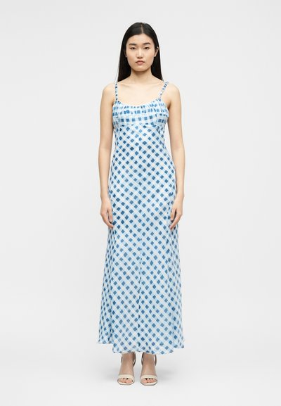 Νεαρή γυναίκα φοράει αμάνικο μακρύ φόρεμα με μπλε και λευκά καρό και μπεζ ανοιχτά παπούτσια, στέκεται μπροστά σε απλό λευκό φόντο.