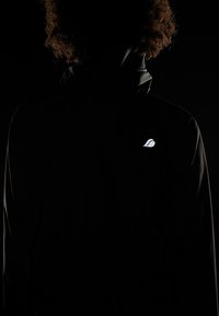 Veste noire à col montant, fabriquée dans un matériau lisse et léger. Présente un logo réfléchissant dans le dos, créant un contraste subtil.