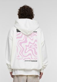 Felpa bianca con cappuccio caratterizzata da un motivo ondulato rosa sulla schiena, con testo nero sopra e sotto il design. Tessuto morbido, vestibilità comoda.