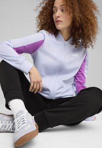 Svetlo modra zip-jakna z vijoličnimi detajli, v kombinaciji s črnimi hlačami. Bele nogavice in svetlo sivi športni copati z gumijastimi podplati dopolnjujejo obleko.