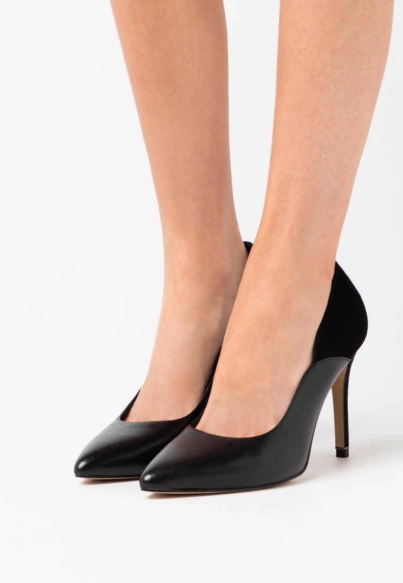 Jambes portant des escarpins noirs à bout pointu avec talons aiguilles sur un fond blanc.