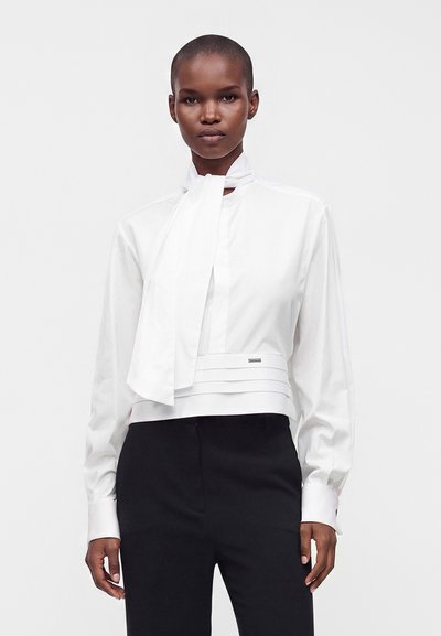 Femme portant un chemisier blanc à manches longues avec un détail nœud au col et un pantalon noir taille haute, debout devant un fond uni.