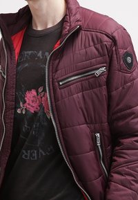 Kaštanová zateplená bunda s prošívanou texturou, která má černou kapsu na zip a červenou vnitřní podšívku. Nošena přes černé tričko s potiskem.