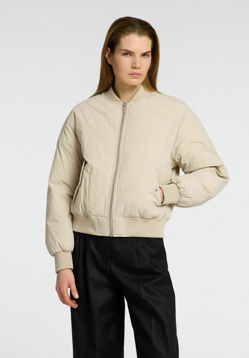 Una donna con capelli lunghi indossa una giacca bomber beige e pantaloni neri, in piedi con sicurezza con una mano in tasca, su uno sfondo neutro.