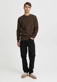 Brun teksturert genser, svarte bukser og grå joggesko med hvite striper. Modell poserer med den ene hånden i lommen, nøytral bakgrunn.