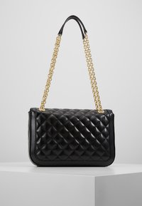 Svart kviltad läderhandväska med guld dubbelkedjeslinga, uppställd på en vit pedestal mot en enkel grå bakgrund.