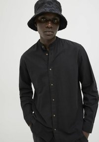 Camicia nera a maniche lunghe con colletto alla coreana, caratterizzata da bottoni marroni e un cappello a secchiello testurizzato, entrambi coordinati in nero.