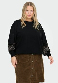 Melna džemperis ar rakstainām, tekstūrētām piedurknēm, valkāts virs brūnas korduroja svārku, kam ir priekšējie pogas un kabatas.