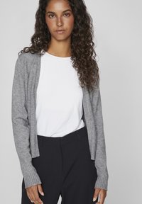 Grijze gebreide cardigan met ribbels aan de mouwen, gedragen over een witte t-shirt, gecombineerd met marineblauwe broek. Ontspannen pasvorm en zachte textuur.