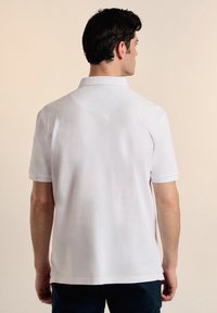 Camisa polo blanca hecha de una tela suave y transpirable, que presenta un cuello clásico y mangas cortas, con un dobladillo recto y sutiles aberturas laterales.