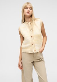 Νεαρή γυναίκα με ξανθά μαλλιά, φορούσα αμάνικο πλεκτό κίτρινο γιλέκο με κουμπιά και μπεζ παντελόνι, με το χέρι στην τσέπη, να στέκεται μπροστά σε λευκό φόντο.