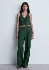 Un gilet court à rayures vertes avec trois boutons associé à un pantalon ample taille haute. Le tissu est d'une texture lisse. Design minimaliste.