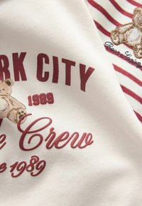 Valkoinen kangas, jossa on suuri burgundinvärinen graafinen teksti "NEW YORK CITY" ja "1989." Mukana myös nallekarhun kuva ja raidalliset yksityiskohdat.