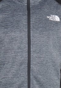 Grå zip-jacka gjord av ett texturerat tyg. Har en svart dragkedja och en vit logotyp på den övre vänstra bröstområdet.