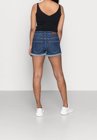 Kvinna i en svart  ärmlös tröja, uppvikta denimshorts och vita sneakers som står vänd mot en vit vägg.