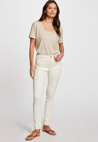 Top beige de manga corta con escote redondeado, combinado con pantalones ajustados crema. Sandalias planas marrones completan el atuendo. Texturas suaves y diseño ajustado.