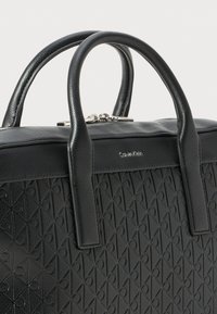 Bolso negro con un diseño texturizado que presenta patrones de diamantes y corazones. Hecho de material sintético con mangos resistentes y un cierre de cremallera plateado.
