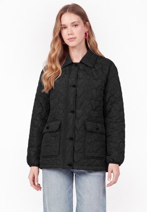 Femme aux longs cheveux blonds portant une veste matelassée noire avec des motifs de cœurs, boutonnée à l'avant, avec des poches à rabat, et un jean bleu clair.