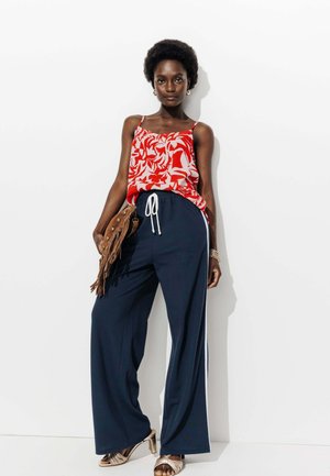 Débardeur à fleurs rouges avec des fines bretelles, associé à un pantalon large marine avec des rayures blanches sur les côtés. Sac à franges marron et sandales à talons métalliques.