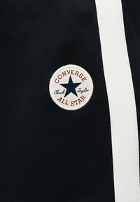 Στρογγυλή ετικέτα λογότυπου Converse All Star με ναυτικό αστέρι και κείμενο σε μαύρο ύφασμα δίπλα σε κάθετη λευκή ρίγα.