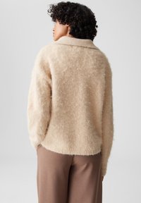 Maglione beige morbido con colletto a costine, texture soffice e vestibilità ampia. Indossato sopra pantaloni marroni, caratterizzati da linee semplici e pulite.