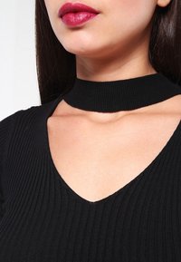 Nainen, jolla on tummat suorat hiukset, pukeutuneena mustaan ribbitoppiin, jossa on choker-kaulus, ja tummanpunaista huulipunaa, yksinkertaista taustaa vasten.