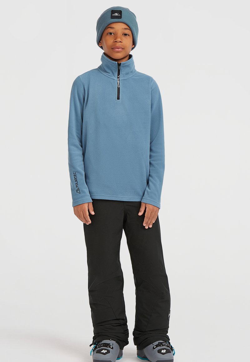 Blå fleecepullover med lynlås i kraven, parret med sorte snebukser og grå sneboot med turkise detaljer.