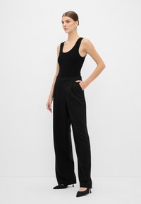 Zwarte geribbelde body gecombineerd met hooggeplaatste zwarte broek met streepjes; model staat met een hand in de zak, draagt puntige zwarte hakken.