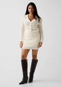 Ensemble cardigan tricoté et jupe crème, avec tissu texturé et boutons dorés. Le modèle porte de hautes bottes en cuir marron.