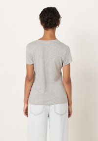 Oseba z kodrastimi lasmi v figi, oblečena v svetlo sivo majico s kratkimi rokavi in svetlo modre kavbojke, obrnjena stran s preprostim ozadjem.