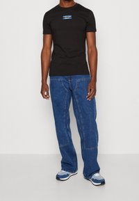 Black cotton t-shirt with blue logo, paired with light blue denim jeans featuring a loose fit and visible stitching. Blue and white sneakers complete the look.