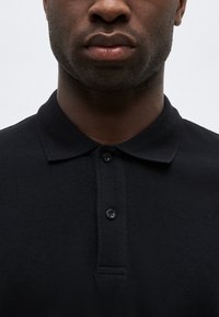 Κοντινό πλάνο ενός ατόμου που φοράει μαύρο πόλο πουκάμισο με κουμπιά, δείχνοντας το κάτω μισό του προσώπου και το λαιμό σε απλό φόντο.