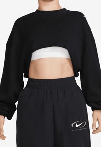 Svart kort sweatshirt med en urtagning i mitten, kombinerad med en vit behå och svarta träningsoveraller med en silverfärgad Nike-logga. Mjuk, avslappnad passform.