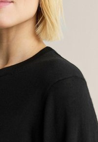 Svart stickad tröja med rund halsringning, visar en mjuk textur och lätt drapering. Närbild som framhäver kragen och axeln.