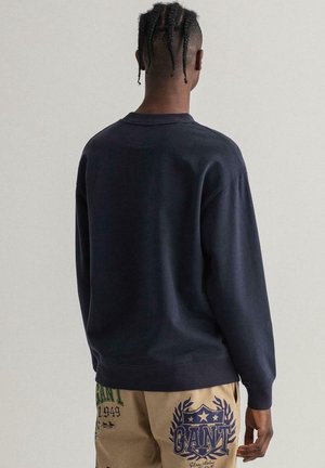 Homme aux cheveux tressés portant un sweat-shirt bleu marine foncé et un pantalon beige avec de grands logos imprimés en bleu marine et vert, debout devant un fond uni.