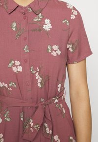 Sukienka z guzikami w stonowanym różowym kolorze, z kwiatowym wzorem i motylami, krótkimi rękawami oraz talią wiązaną w detalu.