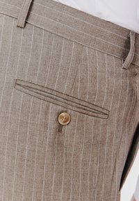 Jasnobrązowe spodnie w prążki mają tylną kieszeń z pojedynczym zapięciem na guzik oraz gładki, teksturowany materiał.