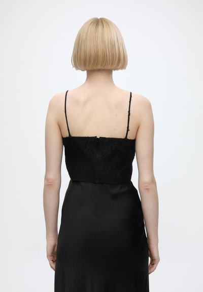 Personne blonde avec une coiffure courte au carré portant une robe noire sans manches avec des bretelles fines, vue de dos sur un fond uni.