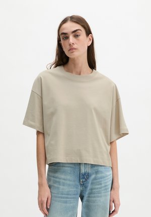 Jeune femme aux longs cheveux bruns portant un haut beige oversize à manches courtes et un jean bleu clair, debout sur un fond blanc uni.