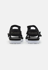 Zapatos tipo sandalia negros con correas ajustables y una plantilla texturizada, que presentan una suela blanca con una banda de acento negra y un logo en el talón.
