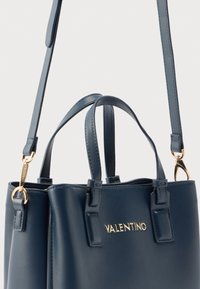 Τσάντα χειρός από δέρμα σε ναυτικό μπλε με χρυσά μεταλλικά στοιχεία, κοντές λαβές και αποσπώμενο λουράκι. Διαθέτει λογότυπο και δομημένο σχέδιο. Με ομαλή υφή.