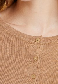 Braunes, geripptes Strickoberteil mit einem runden Halsausschnitt und drei dekorativen beigen Knöpfen an der Vorderseite. Weicher Stoff und taillierte Passform.