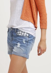 Deminshorts i ljusblått med slitna detaljer, uppvikta fållar och blommönsterbroderi på bakfickan. I kombination med en vit topp och en orange cardigan.