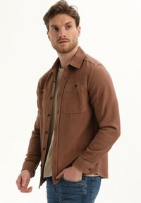 Rjava srajca s gumbi iz teksturirane tkanine, z dvema žepoma na prsih, dolgimi rokavi in sproščenim krojem. Oblečena čez bež top.
