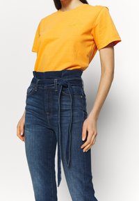 Camiseta de algodón naranja de manga corta con logo amarillo en el pecho, combinada con unos vaqueros de mezclilla oscura de cintura alta que llevan un cinturón atado.